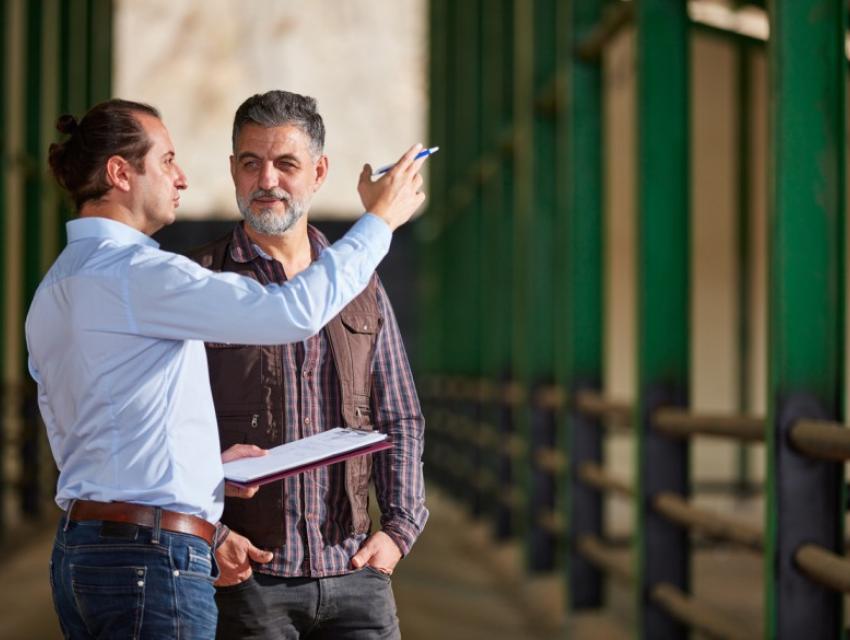 Two men, one pointing with a pen and holding a clipboard, standing and discussing inside a barn with green pillars.