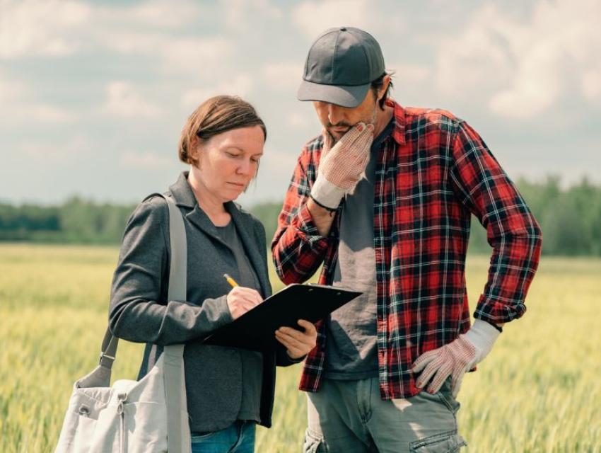 Woman in a gray jacket writing on a clipboard and man in a red plaid shirt with gloves standing in a green field under a cloudy sky.