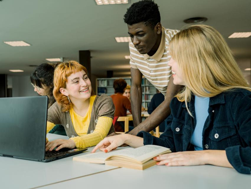 Three students collaborating around a laptop and open book at a table in a library.