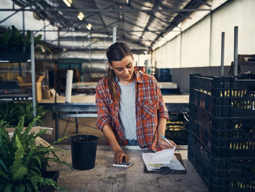 Woman in a plaid shirt using a calculator and clipboard at a table with plants and crates in a greenhouse.